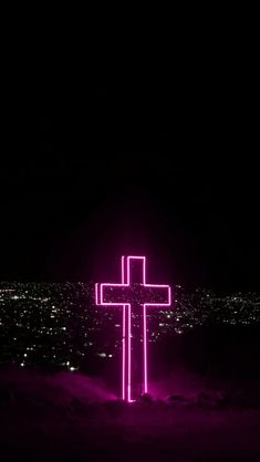 a cross lit up in the dark with purple light on it's side and city lights behind it