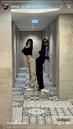 two women are taking selfies in the mirror at an office building hallway with tiled floors and walls