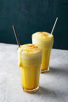 two glasses filled with yellow liquid and topped with whipped cream, on a white surface