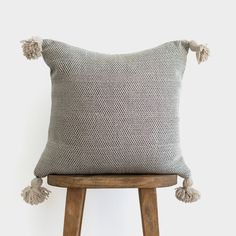 a grey and white pillow with tassels on the back, sitting on a wooden stool