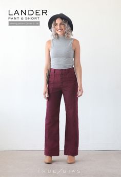 a woman standing in front of a white wall wearing purple pants and a black hat