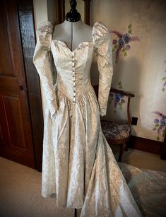 This renaissance-style dress is truly unique. The fitted princess line bodice has a square neckline front and back with a small vee at center front. This is accentuated by a mock opening in the front with "pearl" buttons, each encircled with rhinestones. Some of the buttons are missing a rhinestone. Puff sleeves, with elastic at the shoulders for wearing ease, taper to points at the wrist with a four button and loop closure. The drop waist come to a point in the front and back. Skirt is gathered. Zipper closure. What makes this truly renaissance looking is the fabric. It has gold metallic slightly raised flowers on a cream background. Dress has been cleaned and preserved since the 1991s and is in excellent condition.  Not only would this make a wonderful wedding dress, it would also be ama Elegant Gown With Fitted Bodice And Heart-shaped Neckline, Formal Gown With Fitted Bodice And Sweetheart Neckline, Fitted Ball Gown With Ruched Bodice And Sweetheart Neckline, Elegant Corset Dress With Boned Bodice For Debutante Ball, Fitted Ball Gown With Ruched Bodice, Formal Gown With Sweetheart Neckline And Fitted Bodice, Heart-shaped Neckline Gown With Fitted Bodice For Debutante Ball, Fitted Corset Dress With Boned Bodice And Empire Waist, Fitted Empire Waist Corset Dress With Boned Bodice