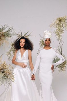 two women in white dresses holding hands and standing next to each other with plants behind them