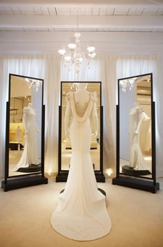 a white wedding dress is displayed in front of three large mirrors with lights on them