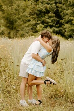 two people are hugging in a field with tall grass and trees behind them, one person has her arms around the other's neck