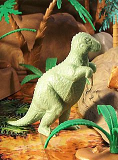 a toy dinosaur is on the ground in front of some fake palm trees and rocks