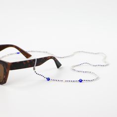 CANDIDASA EVIL EYE SUNGLASSES CORD  Introducing the Candidasa Evil Eye Sunglasses Cord, a dainty and subtle design with classic deep blue evil eye charms. Evil eye symbols have been found for thousands of years across many cultures, they are said to ward off evil, bring positive energy, and even good luck!  - Made with blue glass seed beads, shimmering troca shell pearlised beads, glass evil eye charms, freshwater pearls, wax polyester cord, rubber loops and silver-plated copper alloy findings. Beaded Clear Glass Glasses Chains, Clear Beaded Glass Glasses Chains, White Glass Fashion Accessory Glasses Chains, White Glass Glasses Chains For Fashion Accessory, White Glass Glasses Chains, Trendy White Glasses Chains For Festival, Silver Beaded Glasses Chains For Beach, Trendy Blue Glass Glasses Chains, Silver Glass Glasses Chains For Summer