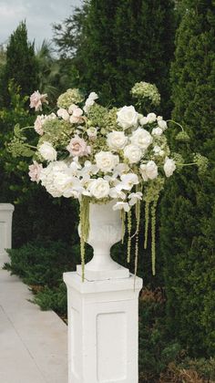 a white pedestal with flowers in it on the side of a road next to bushes