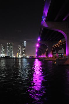 purple lights shine on the water under a bridge