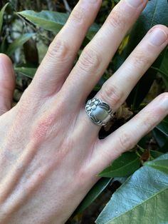 a person's hand with a silver ring on top of it, surrounded by leaves