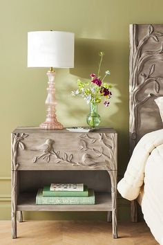 a nightstand with flowers and books on it next to a bed in a green room