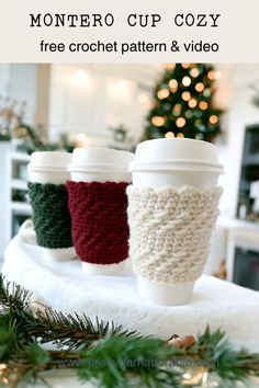 three knitted coffee cups sitting on top of a white blanket next to a christmas tree