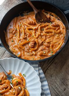 a pan filled with pasta and sauce on top of a wooden table next to a white plate