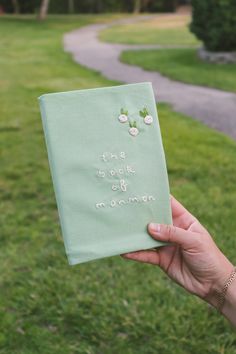 a person holding up a book with embroidered words on it in front of a path