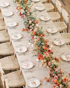 a long table is set with white plates and flowers on the runner, along with place settings