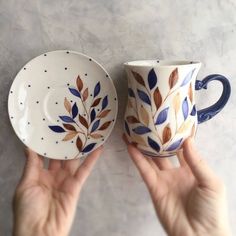 two hand painted coffee cups and saucers with leaves on them, one being held by the other