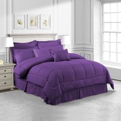 a bed with purple comforter and pillows in a white room next to a window