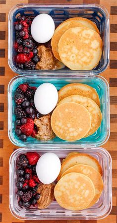 two plastic containers filled with pancakes, berries, and eggs sitting on top of a wooden table