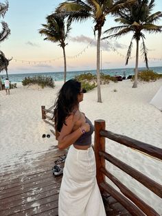 a woman in a white skirt standing on a wooden deck next to the ocean and palm trees