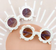 two white sunglasses sitting on top of a pink surface with sun glasses in the background