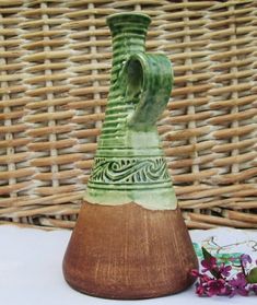 a green and brown vase sitting on top of a white table next to purple flowers