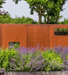 an orange wall with purple flowers in the foreground and trees in the back ground