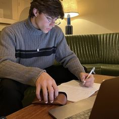 a young man sitting at a table with a laptop and pen in his hand while writing on paper