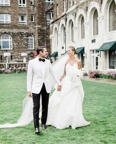 a bride and groom holding hands walking through the grass in front of an old building