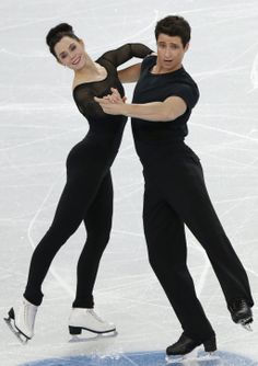 two people skating on an ice rink in black outfits and white shoes, one is holding his arm around the other's shoulder