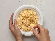 two hands holding a spoon over a bowl of shredded cheese on a white table top