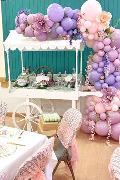 a table topped with lots of balloons next to a white cart filled with pink and purple flowers