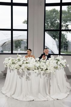 a bride and groom sitting at a table with white flowers in front of large windows