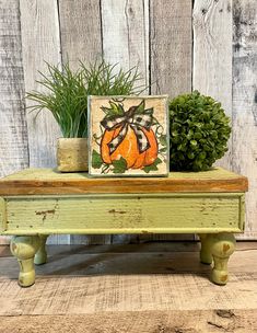 a small wooden bench with a painting on it and some potted plants next to it