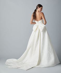 a woman in a white wedding dress with a big bow on her back, standing against a gray background