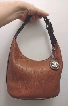 This Dooney & Bourke handbag is a stylish accessory for any occasion. The bag is made of brown caramel leather with a pebbled finish and features a zip-around closure, top handle, and adjustable strap. It is a small hobo size bag and measures 10 inches in width, 6 inches in height, and 5-3/4 inches in depth and has a key holder and bag charm. It is in very good condition with only one small stain on the outer portion of the bag as shown in the photos and some minor slight signs of wear on the interior lining. There is some peeling on the sides of the strap where the bonding is but not on the top or bottom of the strap. It is not noticeable when using. The interior of the bag is lined with beige fabric and has inner pockets for storage. The bag is stain-resistant and water-resistant because Hobo Purses Dooney & Bourke, Chestnut Brown Color, Hobo Bags, Dooney Bourke Handbags, Hobo Handbags, Shoulder Handbag, Leather Hobo, Handbag Purse, Key Holder