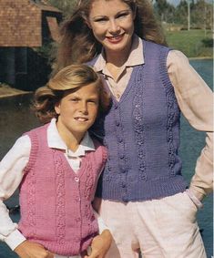 two women in sweaters and vests standing next to each other near the water