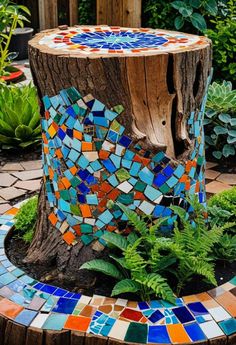 a tree stump with colorful tiles on it and plants around the base in a garden