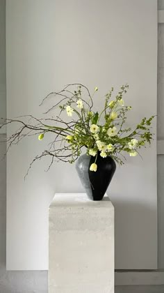 a black vase with yellow flowers in it sitting on a white pedestal next to a wall