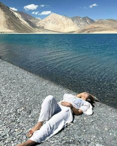 a woman laying on the ground next to a body of water with mountains in the background