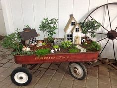 a red wagon filled with plants next to a white house on top of a brick floor