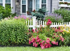 a white picket fence surrounded by colorful flowers and greenery in front of a house