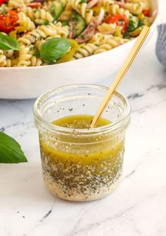 pasta salad with pesto dressing in a glass jar on a marble countertop next to a white bowl and wooden spoon