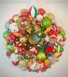 a christmas ornament wreath hanging on the front door with ornaments around it and an elf figurine