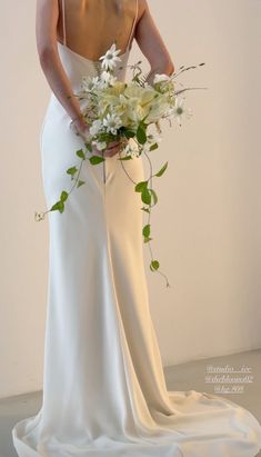 a woman in a white wedding dress holding a bouquet of flowers and greenery with her back to the camera