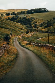 an empty road in the middle of a grassy field with hills and trees on either side