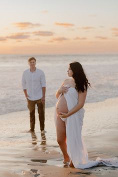 a pregnant woman is standing on the beach with her husband in the background at sunset