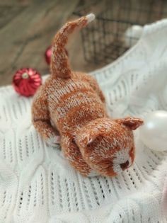 a small stuffed animal cat laying on top of a white knitted blanket next to a red ornament