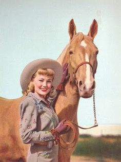 a woman standing next to a brown horse wearing a hat and holding the reigns on her bridle