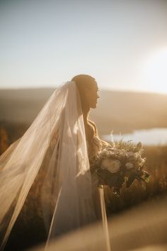 a woman in a wedding dress holding a bouquet and looking at the sun behind her