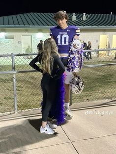 two girls in purple and black cheerleader outfits standing on the side walk looking at each other